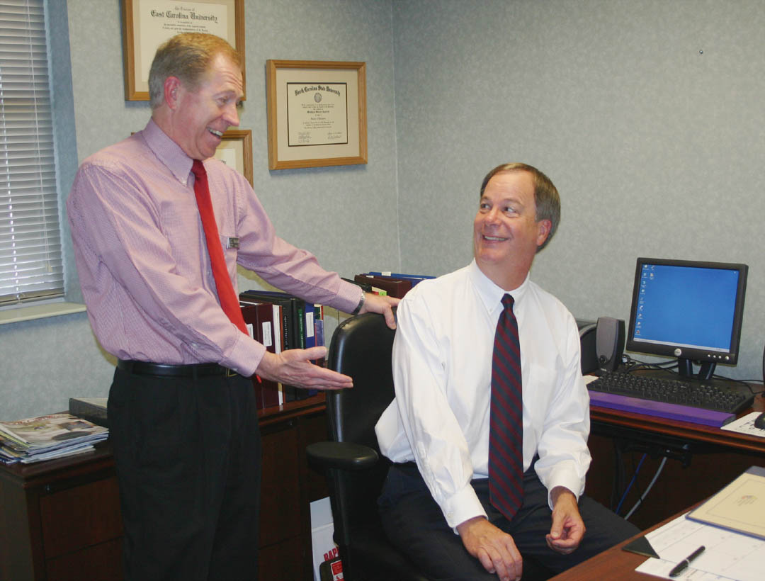 Click to enlarge,  Just-retired Central Carolina Community College President Matt Garrett (standing) and his successor, President Bud Marchant III, share a light moment Aug. 19, as Garrett symbolically welcomes Marchant to the president's chair that Garrett occupied for the past four years. Marchant assumed the presidency on Aug. 15. Garrett's official retirement date is Sept. 1, but he has stepped down from the presidency to be available to assist Dr. Marchant during the transition period for the new president and the college. Marchant is the college's fifth president in its 47-year history.  