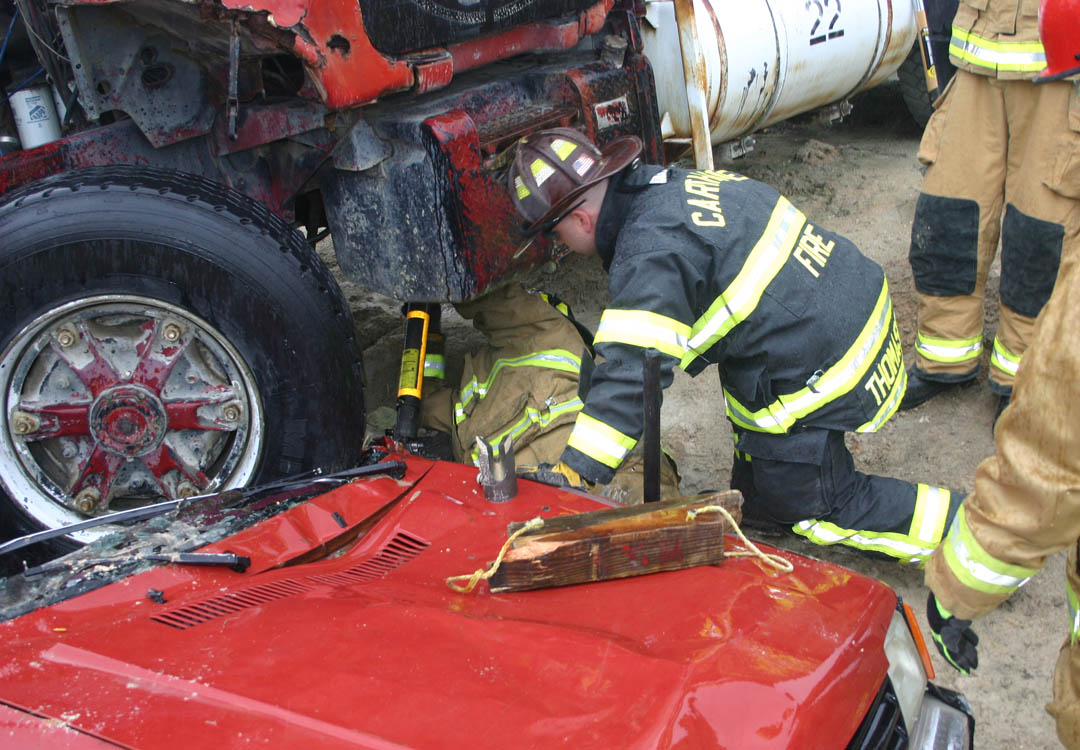 Click to enlarge,  Firefighter Dennis Parker (under truck), of the Noblesville (Indiana) Fire Department, works to set up a stabilizing strut under a concrete mixer truck, assisted by Chad Thomason, of the Carthage Fire Department, as firefighters from other departments observe. After stabilizing it, the firefighters learn how to place lifting straps around it and airbags under it to lift it off the car which is crushed beneath it. The training was part of the Big Rig Rescue scenario held recently at Central Carolina Community College&#8217;s Emergency Services Training Center, in Sanford. Firefighters and rescue squad members came from Virginia, South Carolina and Indiana, as well as nine North Carolina towns and cities, to learn how to safely work with this type of accident. The ESTC not only provides critical training for emergency services personnel, but also boosts the local economy as those coming from out of the area stay at motels, eat at restaurants, and make purchases locally while here. 