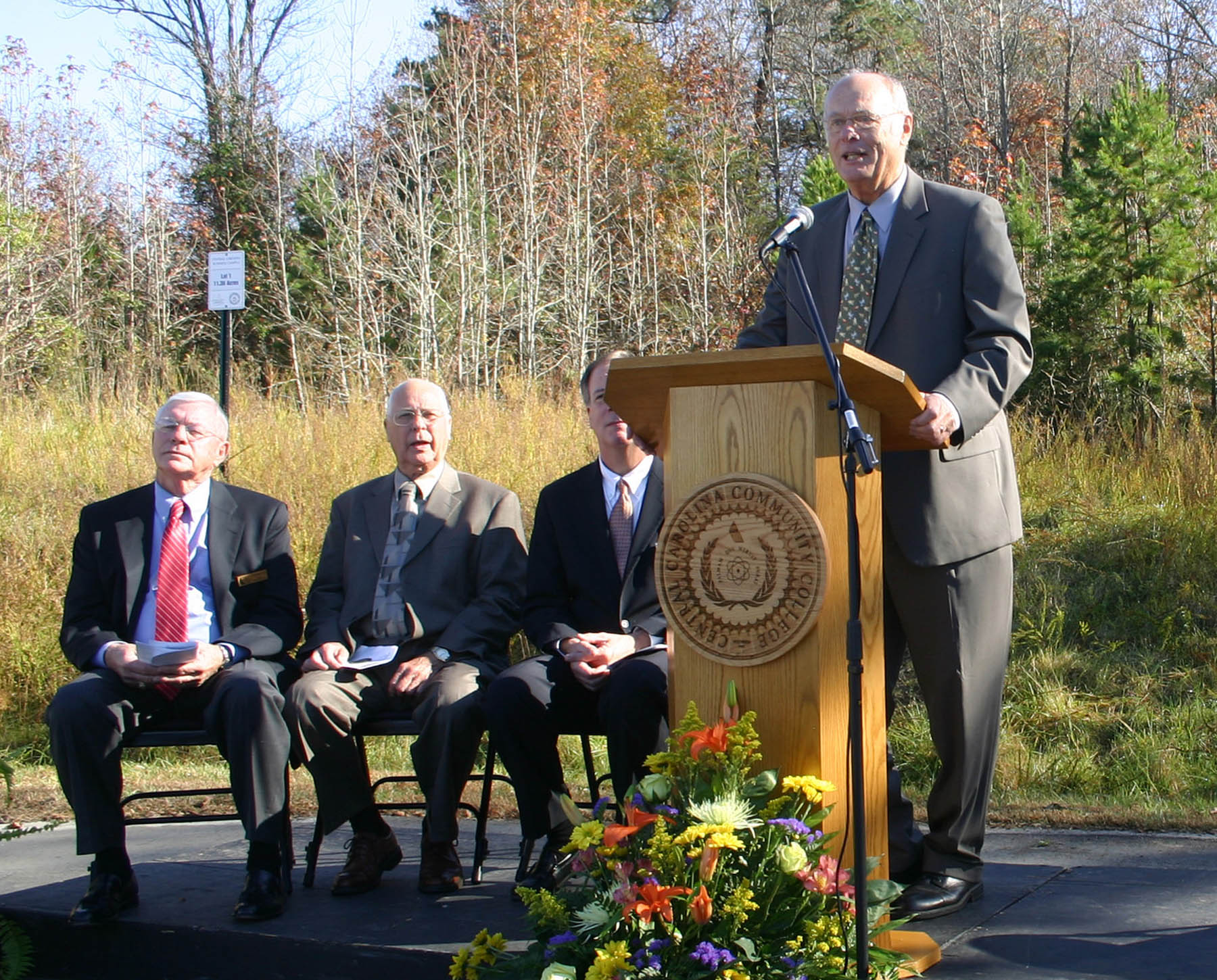 Click to enlarge,  George Lucier, chairman of the Chatham County Board of Commissioners (at podium) addresses those gathered for the groundbreaking ceremony for Central Carolina Community College&#8217;s new Siler City Center. Lucier is also a trustee of the college. The 23,800-square-foot Center, being built in the Central Carolina Business Campus, will enable the college to expand educational opportunities and workforce training for the residents of Siler City and western Chatham County. Also speaking at the groundbreaking were (from left) Bobby Powell, chairman of the Central Carolina Community College Board of Trustees; Charles Turner, mayor of Siler City; Central Carolina Community College President Bud Marchant; and Dianne Reid, president of Chatham County Economic Development Corporation. 