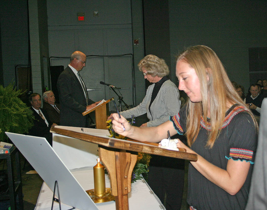 Click to enlarge,  Meredith Albritton, a Central Carolina Community College student from Wilmington, had the honor of signing the membership roll as the first charter member of the college&#8217;s new Beta Sigma Phi Chapter of Phi Theta Kappa International Honor Society. In the background, the second to sign is Sandra Baker, of Dunn. The chartering and membership induction ceremony took place Nov. 4 at the Dennis A. Wicker Civic Center, in Sanford. Fifty-five CCCC students are charter members. Mike Neal (at podium), chapter co-advisor, reads the names of the members as they come forward to sign the charter, as (seated from back left) CCCC President Bud Marchant and college Board of Trustees Chairman Ed Garrison observe. PTK is the largest honor society in American higher education. To be eligible for membership, students must have a minimum of 12 hours of associate degree course work and a grade point average of 3.5 or higher. 