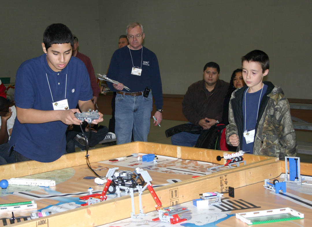 Click to enlarge,  Denis Vielman (left), of West lee Middle School&#8217;s Pride team, controls the team&#8217;s Lego robot while teammate Jacob Marshburn (right) encourages him during the Nov. 20 Central Carolina Community College Robotics Competition at the college&#8217;s Lee County Campus. Observing the action is the competition&#8217;s lead judge Don Hon (standing, center), principal test engineer for EMC Corporation. The West Lee team won the Best Design Award for its robot. The competition, co-sponsored by CCCC, Communities in Schools, and Lee County Schools, was a scrimmage in preparation for the FIRST Lego League Tournament and was conducted by FLL rules. West Lee and SanLee, in Sanford, and two teams from Anne Chestnutt Middle School, in Fayetteville, competed in the scrimmage and will compete in the official 2010 FLL Tournament. The scrimmage and tournament require teams of students to do a presentation on a biomedical problem and then build and program a Lego robot to perform various biomedical tasks, such as inserting a &#8216;stent&#8217; in an &#8216;artery&#8217; made of Legos. 