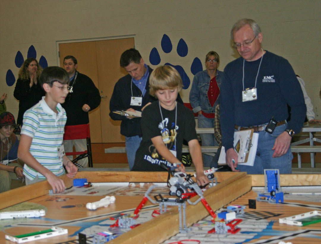 Click to enlarge,  SanLee Middle School student Brandon Donathon (center) works with his team&#8217;s programmed Lego robot at Central Carolina Community College&#8217;s Nov. 20 Robotics Competition at the college&#8217;s Lee County Campus. Teammate Stephen Cameron (left) watches as competition judges John Dills (center, back), of Progress Energy, and Don Hon, principal test engineer for EMC Corporation, take notes. The competition, co-sponsored by CCCC, Communities in Schools, and Lee County Schools, was a scrimmage in preparation for the FIRST Lego League Tournament and was conducted by FLL rules. West Lee and SanLee, in Sanford, and two teams from Anne Chestnutt Middle School, in Fayetteville, competed in the scrimmage and will compete in the official 2010 FLL Tournament. The scrimmage and tournament require teams of students to do a presentation on a biomedical problem and then build and program a Lego robot to perform various biomedical tasks, such as inserting a &#8216;stent&#8217; in an &#8216;artery&#8217; made of Legos. 