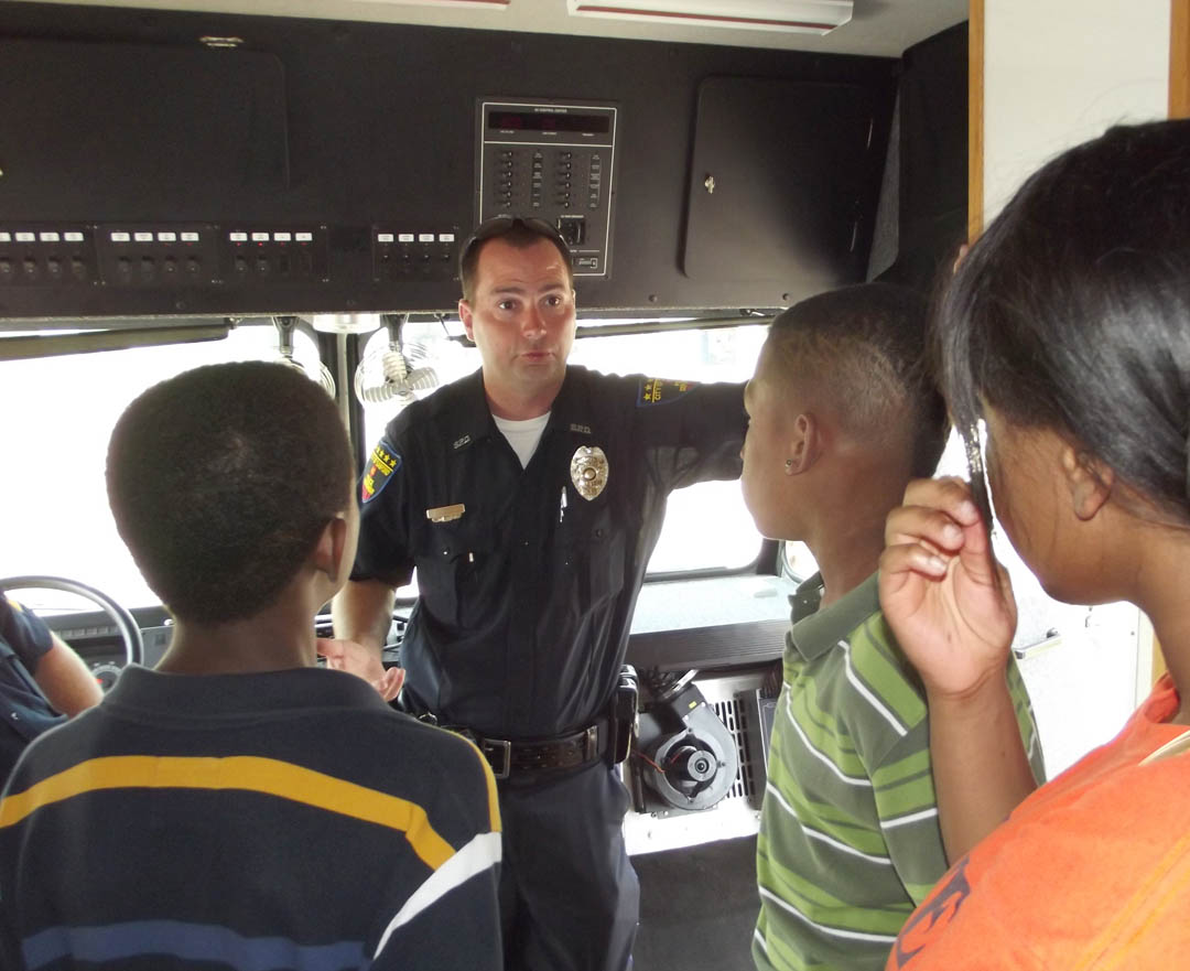 Click to enlarge,  Officer T.W. Herring of the Sanford Police Department explains the department&#8217;s mobile command unit to J.R. Ingram Elementary School fifth graders (from left) DeAndre Brunson and Markeith Watson, as well as Southern Lee High School junior Kierra Harrell. The police department was one of several local agencies and businesses participating Tuesday in Central Carolina Community College&#8217;s Truck-Transportation Day at its Emergency Services Training Center. 