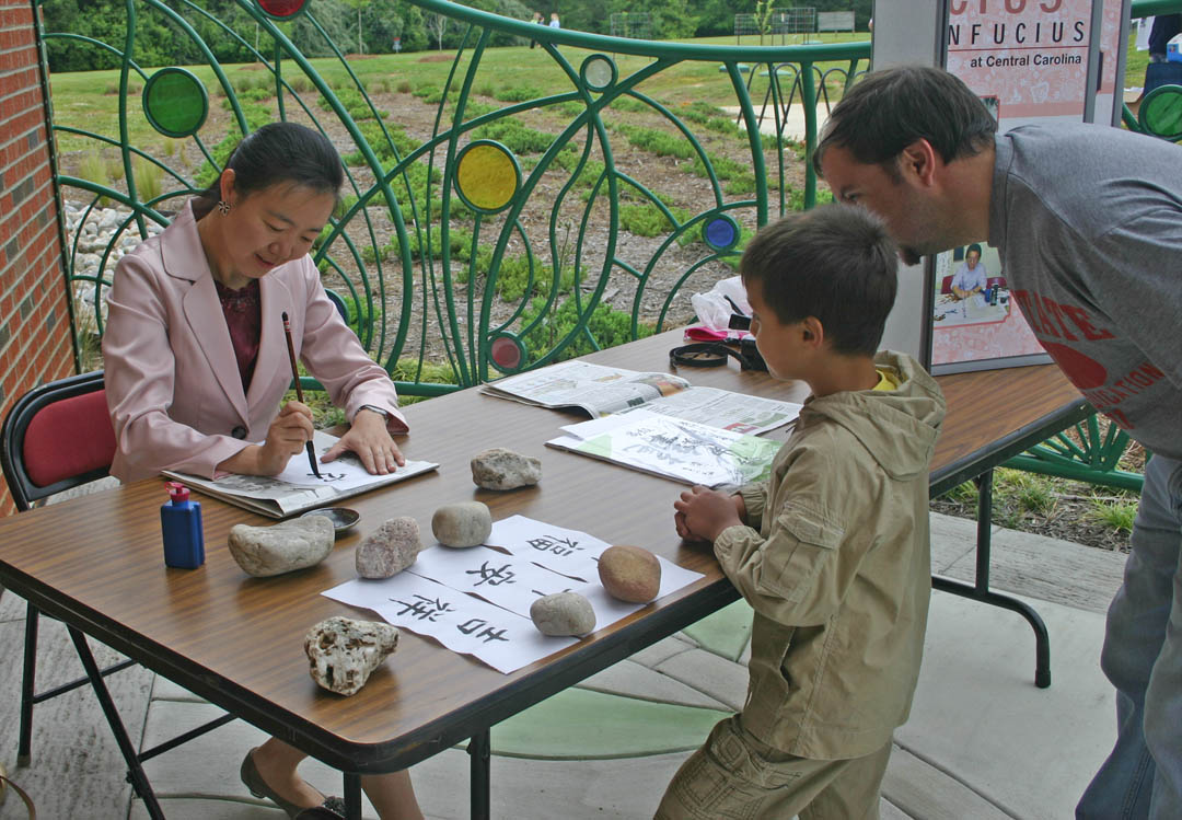 Click to enlarge,  Christopher Watson (right), of Pittsboro, and his son Caden watch with interest as Central Carolina Community College Confucius Classroom instructor Ling Huang (left) draws Chinese words. The calligraphy was a gift to visitors at the college's 50th Anniversary celebration April 28 at the college's Chatham County Campus. It was one of many  attractions at the celebration, which drew hundreds of visitors. The college's 50th celebration will continue, with an event June 2 at the Harnett Campus and a final event June 28 at the Dennis A. Wicker Civic Center. Information about both events will be posted at  www.cccc.edu/50years  as details are finalized. 