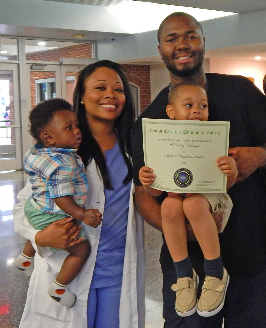 Click to enlarge,  Bridgett Brooks (left) of Harnett County, shares her pride in achievement with her family (from left) son Carter Brooks and husband Eric Brooks, holding son Noah Brooks, after receiving her Phlebotomy Technician certificate during Central Carolina Community College's May 29 Continuing Education Medical Programs Graduation. She plans to continue her education and earn a nursing degree. More than 300 completed their studies during the spring semester in the fields of Nurse Aide I and II, EKG Technician, Phlebotomy Technician, Pharmacy Technician, Central Sterile Processing Technician and Sleep Disorder Technician. Approximately 200 took part in the graduation ceremony. For more information about Continuing Education medical programs, call Lennie Stephenson, 910-814-8833 or email lstephenson@cccc.edu in Harnett County; Heike Johnson, 910-545-8682 or hjohnson@cccc.edu in Chatham County; or Joy McPhail, 919-777-7787 or jmcphail@cccc.edu in Lee County. 