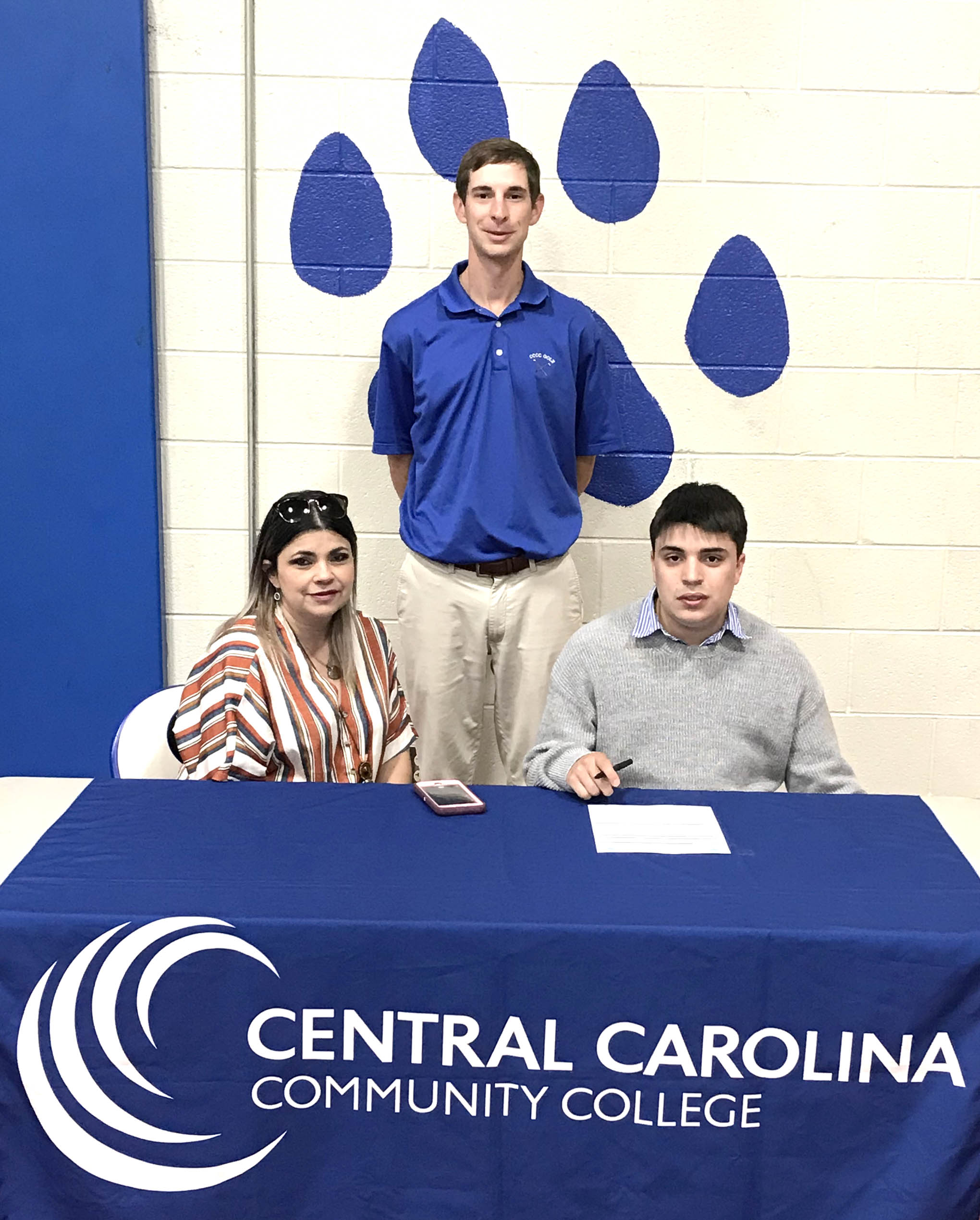 Click to enlarge,  Albanes Figueroa Lugo (seated, right) signs with the Central Carolina Community College golf program as his mother Wanda Lugo join him at the signing. Standing is CCCC Head Golf Coach Jonathan Hockaday. 